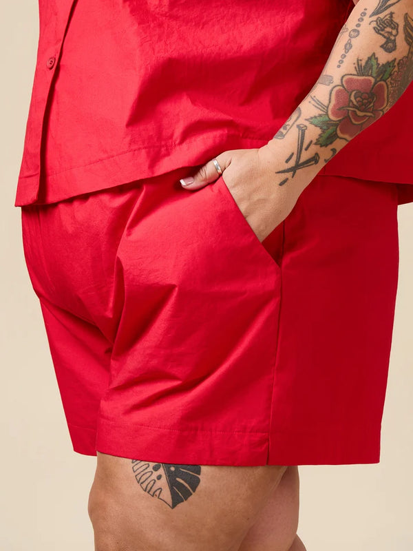 A close-up of a person wearing bright red shorts and a matching red top, with one hand in a pocket and visible arm tattoos, against a plain beige background.
