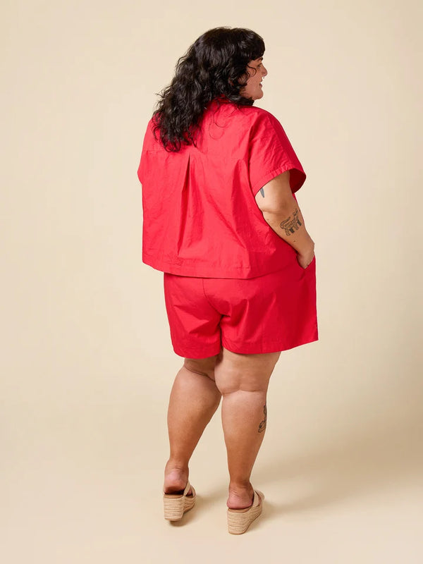 A woman is shown from behind wearing a bright red short-sleeve scrub top and matching red shorts, paired with beige platform sandals, against a solid beige studio background.