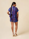 A woman is shown from the back wearing a matching purple two-piece set with a colorful chain print, consisting of a short-sleeve top and shorts, paired with beige espadrille wedge sandals against a solid beige background.