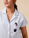 A close-up of a woman wearing a light blue and white vertically striped short-sleeve button-up shirt with a chest pocket holding white sunglasses, against a solid beige background.