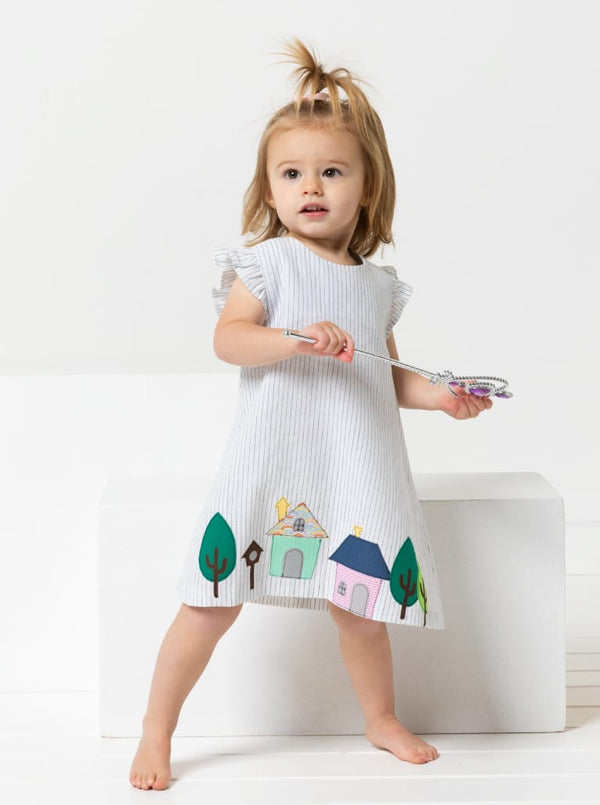 A toddler girl stands barefoot on a white studio set wearing a white ruffled dressmade using andie kids dress sewing pattern with a colorful house and tree print near the hem, holding a toy wand.