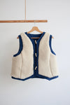 Waistcoat made using Devon Waistcoat Sewing Pattern PDF – Relaxed fit, fully lined, reversible design with patch pockets and button or snap closures. White fleece vest with blue trim on a wooden hanger against a white background