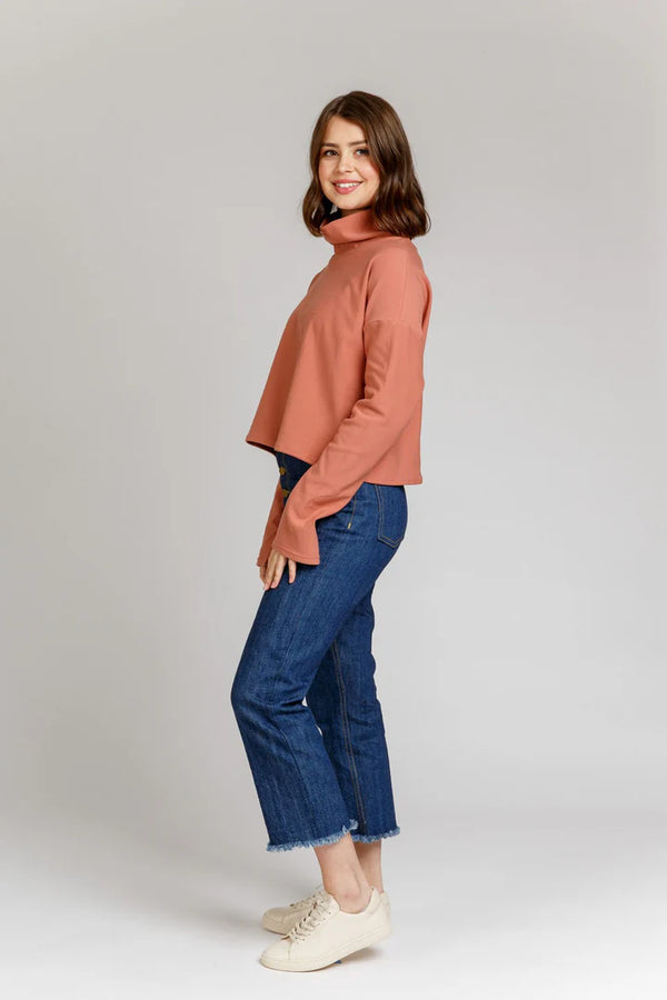 Woman wearing a brown turtleneck Jarrah sweater and blue jeans on a white background