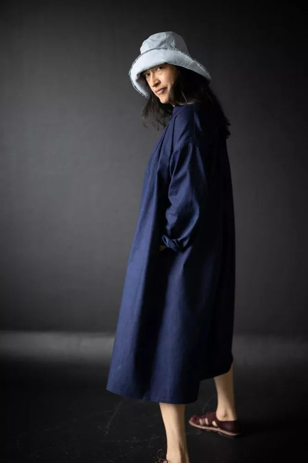 Person wearing a long dark coat and light hat against a dark background