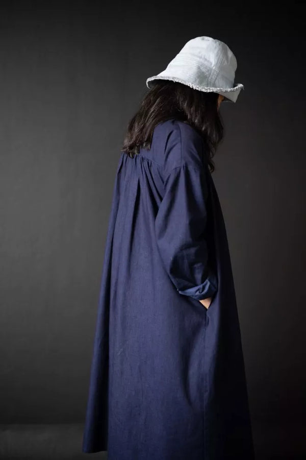 Person wearing a long dark blue The Cawley Sewing Pattern by Merchant & Mills and white bucket hat against a dark background