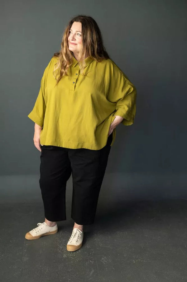 Woman wearing a mustard yellow blouse and black pantsmade using The Eve Trouser Sewing Pattern by Merchant & Mills – cropped tapered trousers with side zip and pleats against a gray background
