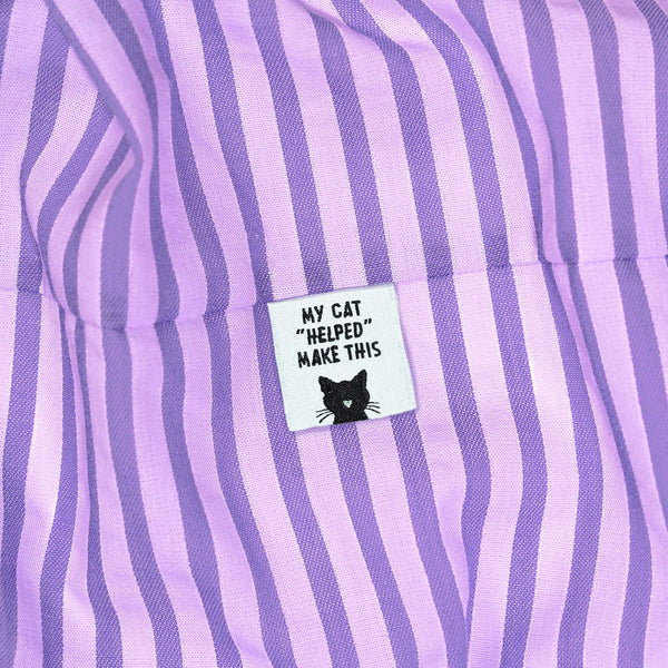 Close-up of a lavender and purple vertically striped fabric item with a small white sewn label that reads “MY CAT ‘HELPED’ MAKE THIS” above a black cat face graphic.