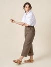 Woman wearing a white shirt and brown pants made of Rika Pants sewing pattern in cropped tailored style with hem cuffs and tabbed waistbandon a beige background