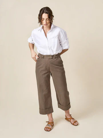 Person wearing a white shirt and brown pants made of Rika Pants sewing pattern in cropped tailored style with hem cuffs and tabbed waistbandon a beigebackground