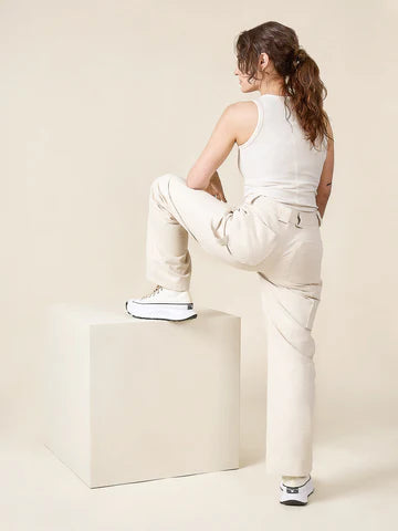 Person sitting on a white cube wearing a white tank top and beige pants against a beige background