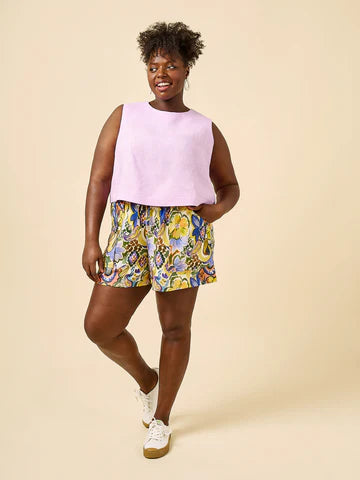 Woman wearing a light purple sleeveless top and colorful floral shortsmade using Sepia Pants and Shorts sewing pattern on a beige background