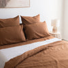 Tobacco brown linen fitted sheet on a bed with a lamp and wicker side table in the background.