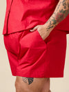 A close-up of a person wearing bright red shorts and a matching red top, with one hand in a pocket and visible arm tattoos, against a plain beige background.
