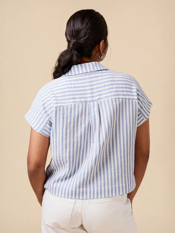 A woman shown from behind wearing a short-sleeve collared blouse with vertical light blue and white stripes, paired with white pants, against a solid beige background.