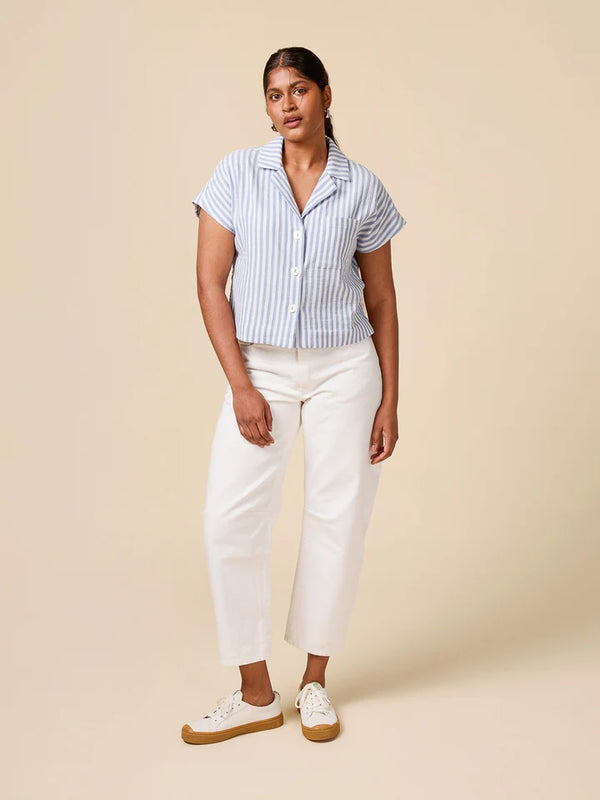 A woman modeling a short-sleeve button-up shirt with light blue and white vertical stripes, paired with white cropped pants and white sneakers with tan soles, against a solid beige studio background.