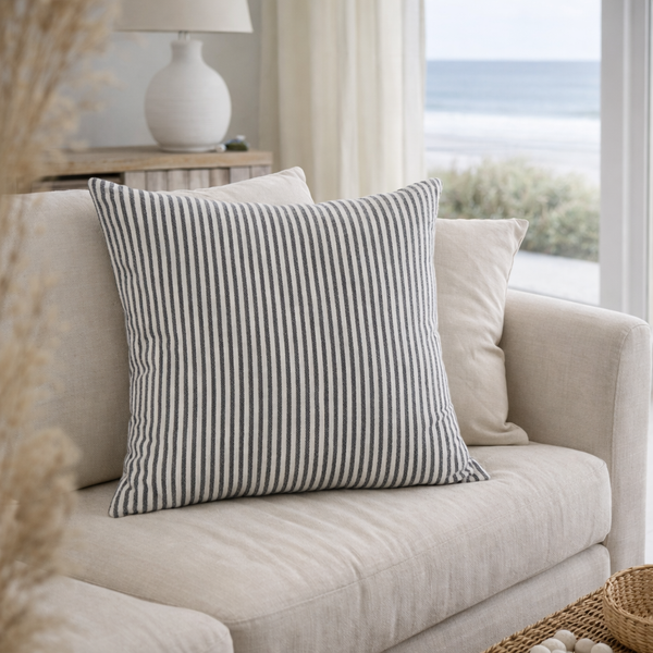 Charcoal Striped pillow on a beige sofa in a living room with a lamp and window in the background.