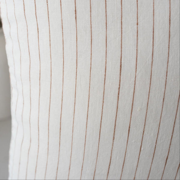 Close-up of a white pillow with brown stripes and a blurred vase in the background.