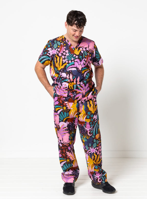 A man wearing a short-sleeve V-neck scrub set with a vibrant, multicolored tropical and coral print, standing against a plain white studio background.