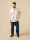 Man wearing a beige jacket, blue jeans, and brown shoes on a beige background