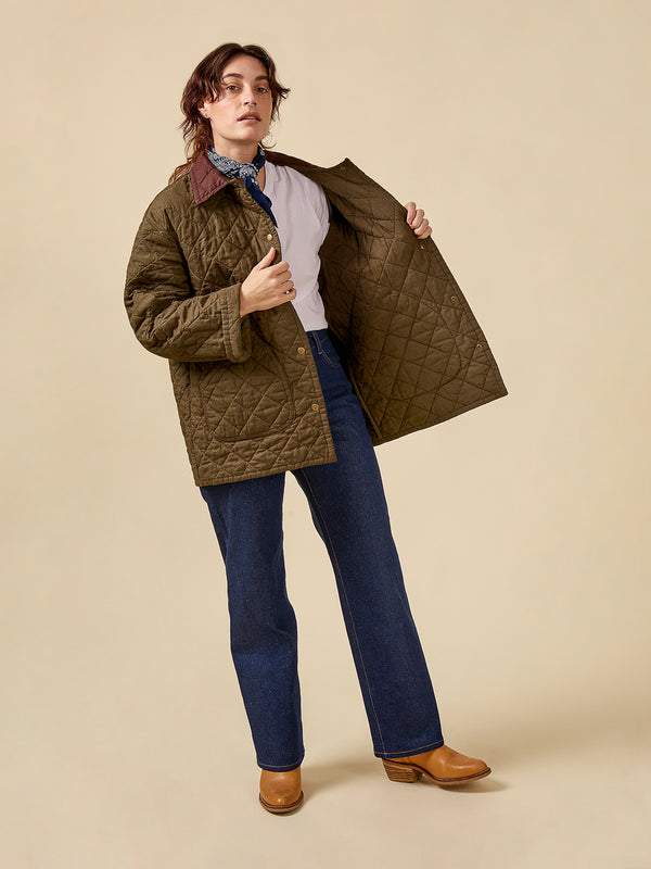 Person wearing a quilted jacket over a white shirt with blue jeans and brown shoes on a beige background