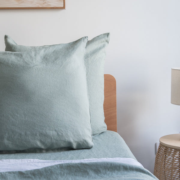 Harbour Sage European linen pillowcaseson a bed with a wooden headboard and lamp in the background.