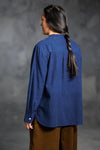 Person wearing a blue top made using The Niven Sewing Pattern by Merchant & Mills – oversized shirt or shirt-dress with bib, grandad collar and cuffs and brown pants against a gray background