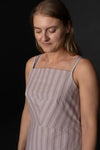 A woman wearing a sleeveless square-neck dress with thin vertical stripes in muted pink, beige, and gray, photographed against a dark studio background.