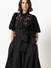 A woman wearing a black lace blouse with short puff sleeves and a high collar, paired with a black skirt made using Sophia Woven Bubble Skirt and Pant sewing pattern featuring a large waist bow, photographed against a plain light background.